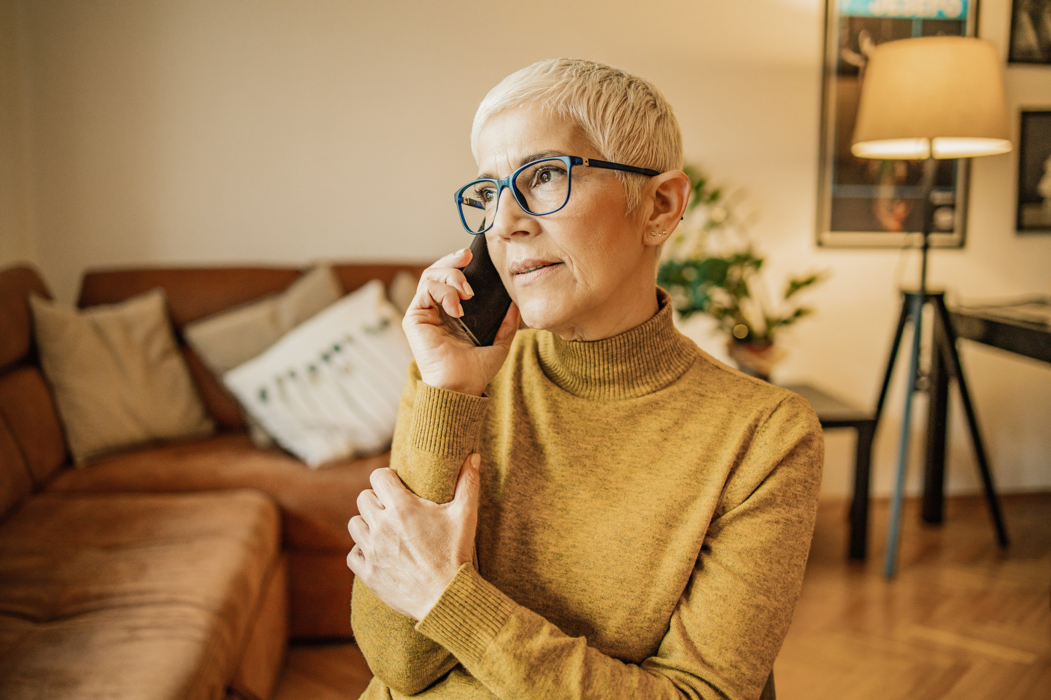 A person on the phone in their living room. They look deeply engaged in the conversation and relaxed. They have thick-rimmed glasses and short, white hair, as well as light skin tone.